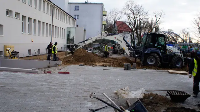 Koparka i robotnicy układają nowy fragment placu przy budynku z zadaszeniem