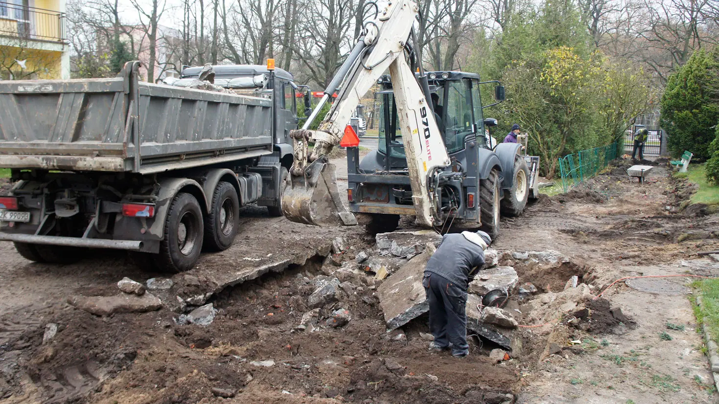Koparko-ładowarka i ciężarówka usuwają rozbity beton, robotnik zbiera gruz z trawnika