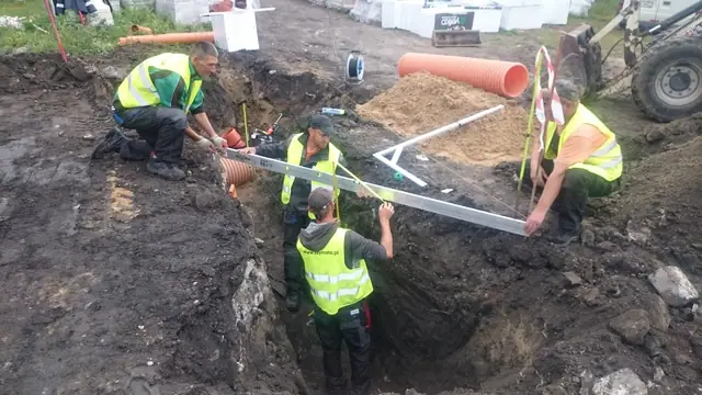Ekipa w kamizelkach odblaskowych opuszcza belkę niwelacyjną do głębokiego wykopu przy budynku