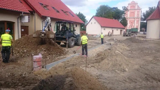 Ekipa robotników i geodeta z niwelatorem pracują na tle wieży kościoła w Skrzatuszu