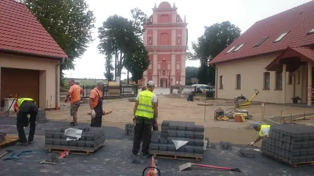 Robotnicy układają ciemną kostkę brukową na dziedzińcu, w tle różowa wieża kościoła