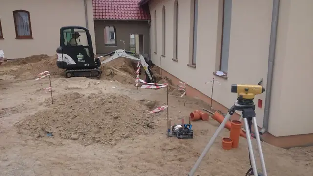 Niwelator na trójnogu ustawiony przy wykopie z rurami kanalizacyjnymi obok mini koparki