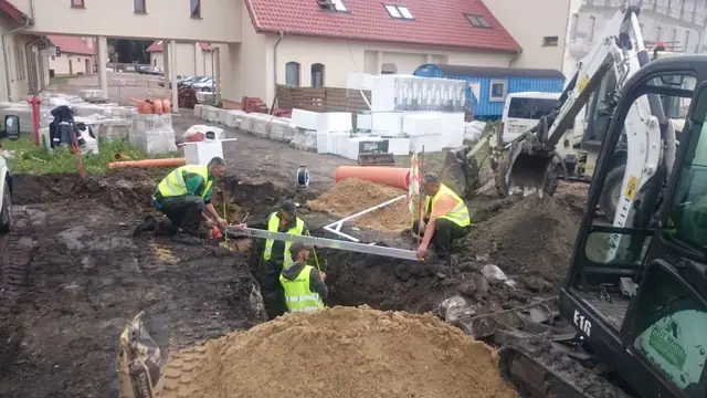 Trzech pracowników w kamizelkach odblaskowych łączy elementy instalacji obok mini koparki