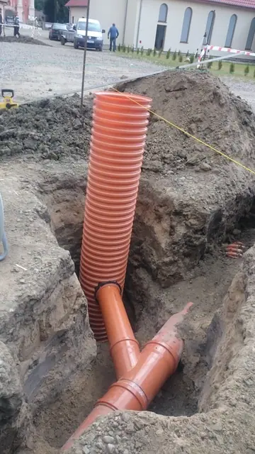 Pomarańczowa rura kanalizacji deszczowej osadzona w wykopie przy ścianie budynku sanktuarium