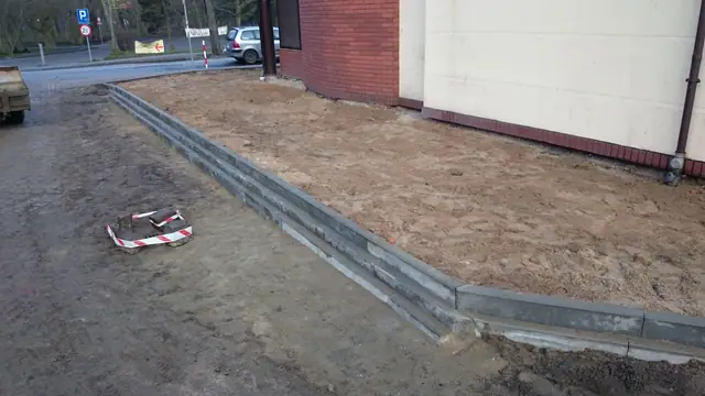 Świeżo osadzony betonowy krawężnik obramowuje rabatę ziemną oznaczoną znakiem ostrzegawczym