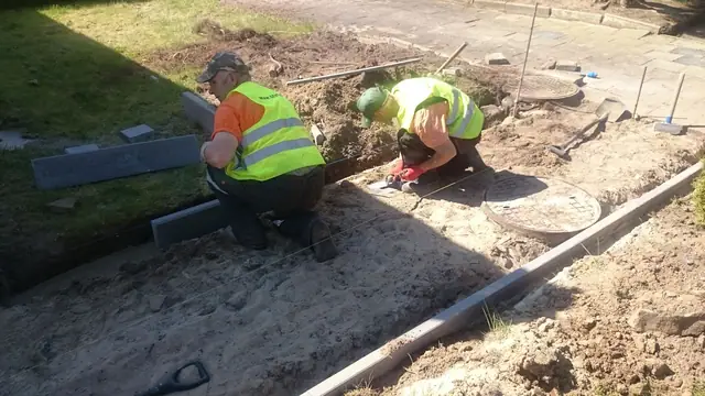 Robotnicy układają krawężnik w wykopie na trawniku podczas wymiany chodnika w ośrodku w Sianożętach