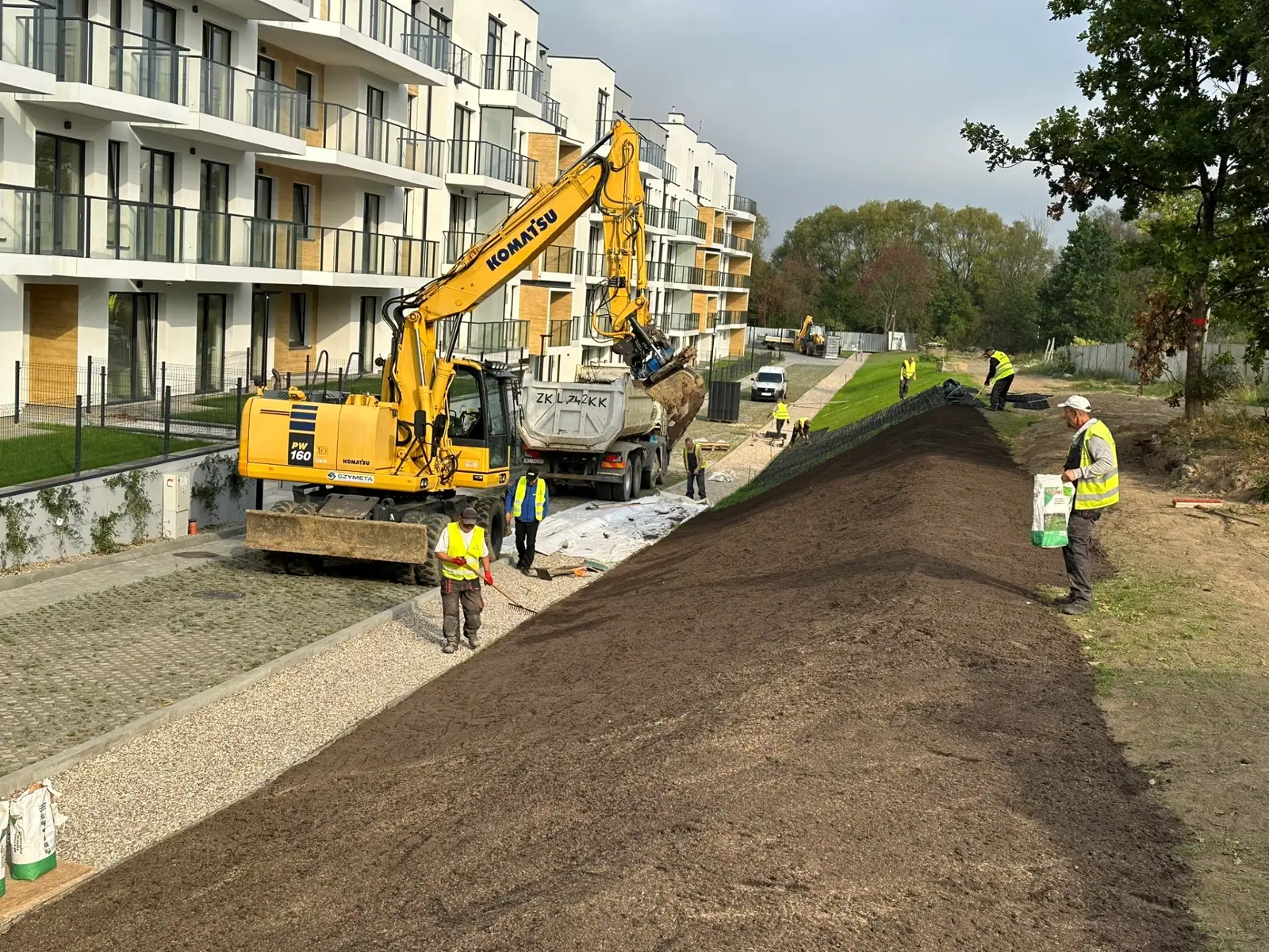 Koparka Komatsu i ciężarówka rozkładają warstwę ziemi na skarpie przy blokach mieszkalnych w Ustroniu Morskim