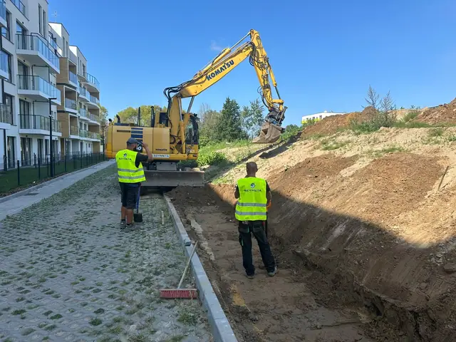 Koparka Komatsu formuje nasyp ziemny wzdłuż chodnika, robotnicy nadzorują prace przy wzmocnieniu skarpy