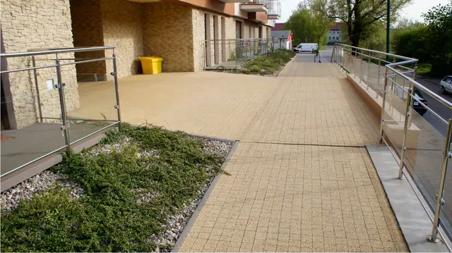 Wybrukowana pochylnia z metalową poręczą prowadząca wzdłuż budynku obok kosza na śmieci
