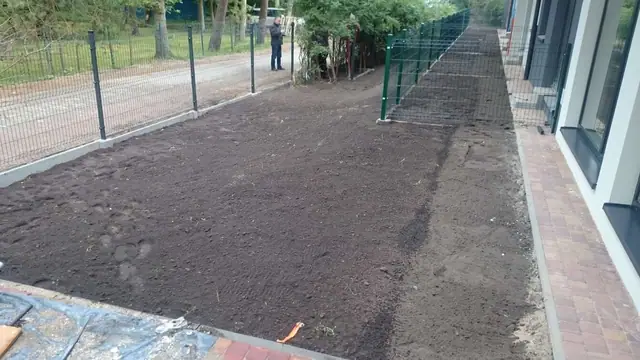 Świeżo rozłożona czarna ziemia wzdłuż siatkowego ogrodzenia obok wybrukowanej alejki