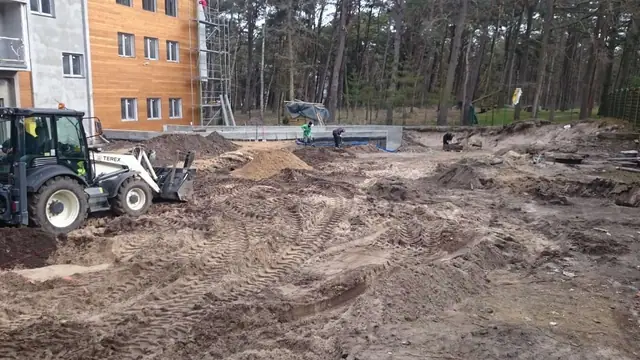 Koparko-ładowarka wyrównuje piaszczysty plac obok drewnianej elewacji budynku