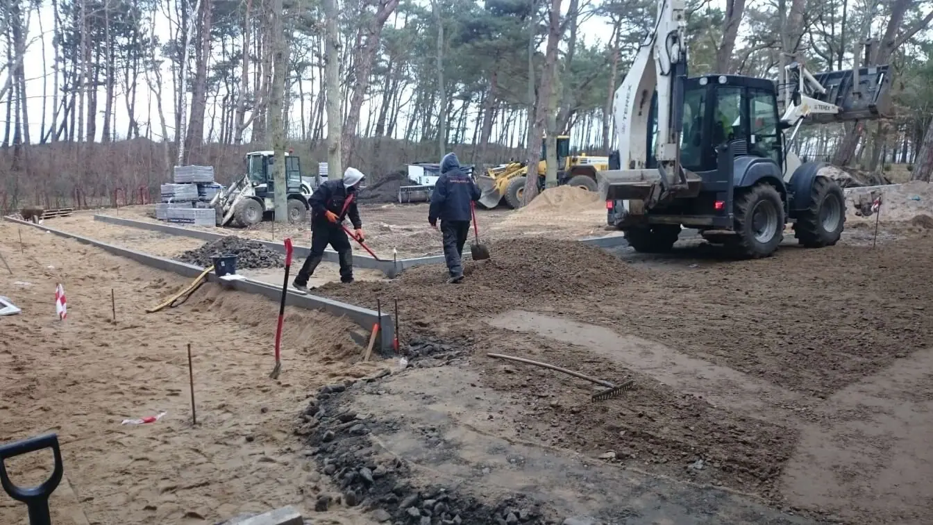 Robotnicy wyznaczają krawężnikami ścieżkę na piaszczystym terenie wśród sosen przy inwestycji Dwie Sosny