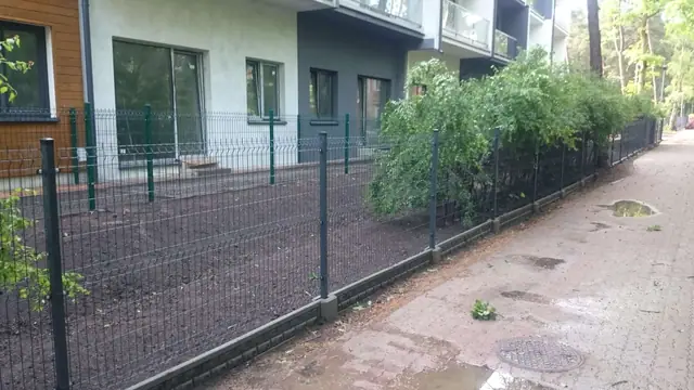 Zielone ogrodzenie siatkowe wzdłuż wybrukowanej ścieżki przy budynku z balkonami