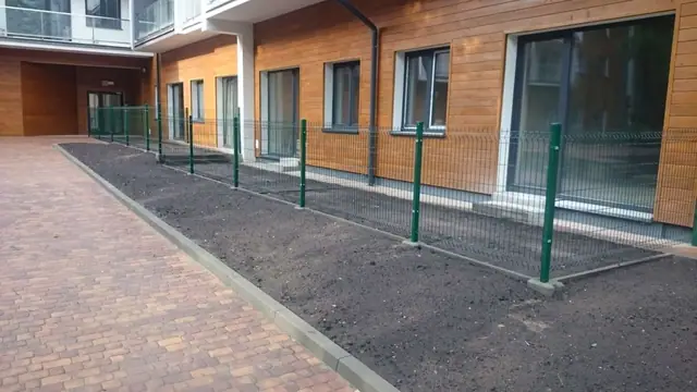 Zielone ogrodzenie z siatki wzdłuż wybrukowanej ścieżki przed wejściem do budynku