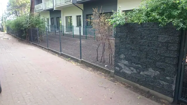 Ścieżka wzdłuż ciemnego muru kamiennego z siatkowym ogrodzeniem przy budynku mieszkalnym