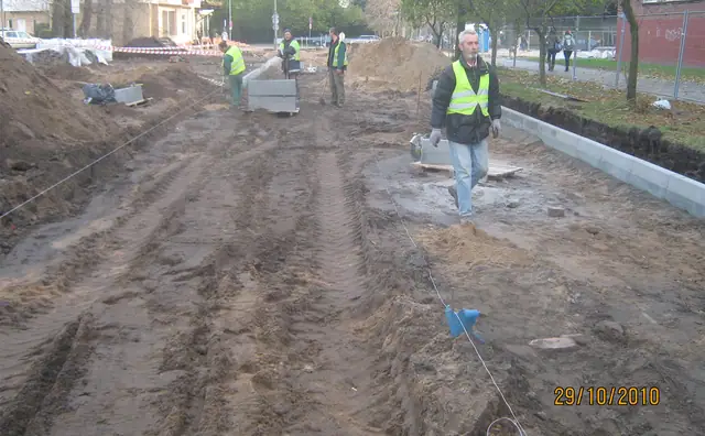 Pracownicy układają krawężnik na piaszczystej alejce kompleksu stadionów