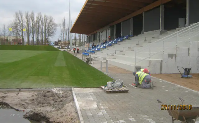 Pracownik zaciera nawierzchnię przy trybunach stadionu z widoczną murawą boiska