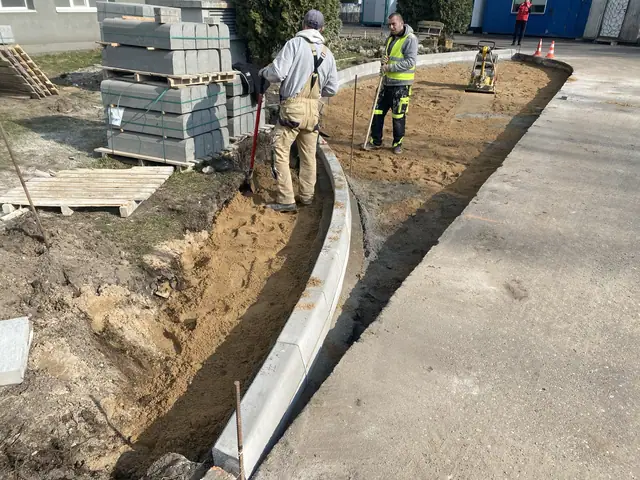 Pracownicy układają łukowy krawężnik betonowy przy wykopie obok materiałów na paletach