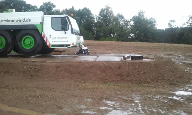 Biało-zielona ciężarówka firmy transportu dźwigów zaparkowana przy platformie fundamentowej turbiny