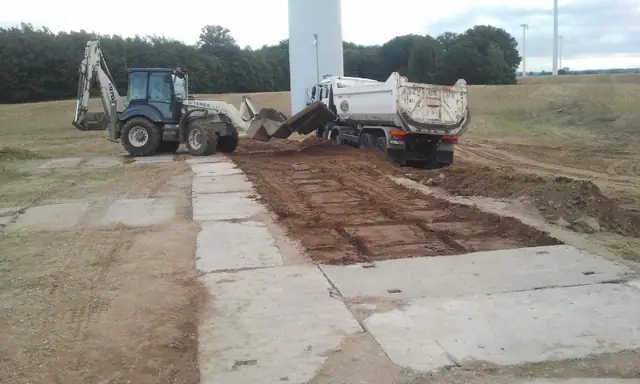 Koparka i wywrotka rozładowują ziemię przy betonowej płycie drogi u podstawy turbiny