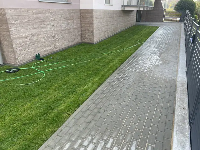 Chodnik z kostki obok trawnika z wężem ogrodowym przy okładzinie kamiennej elewacji