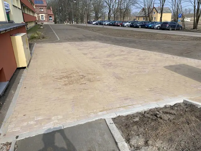 Szeroki plac z beżowej kostki brukowej przed budynkiem z zaparkowanymi samochodami