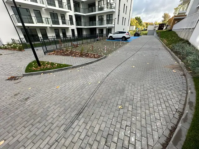 Zakręt drogi wewnętrznej z kostki brukowej wzdłuż elewacji budynku, miejsca postojowe w tle