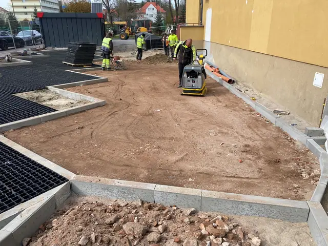 Pracownicy układają krawężniki na piaszczystym podłożu przy budynku mieszkalnym