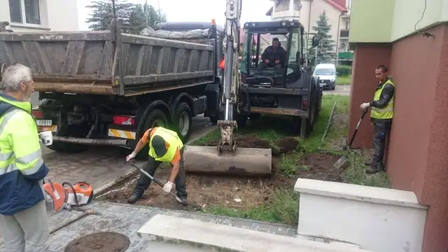 Koparko-ładowarka i robotnicy demontują stary krawężnik przy elewacji budynku, obok ciężarówka z gruzem