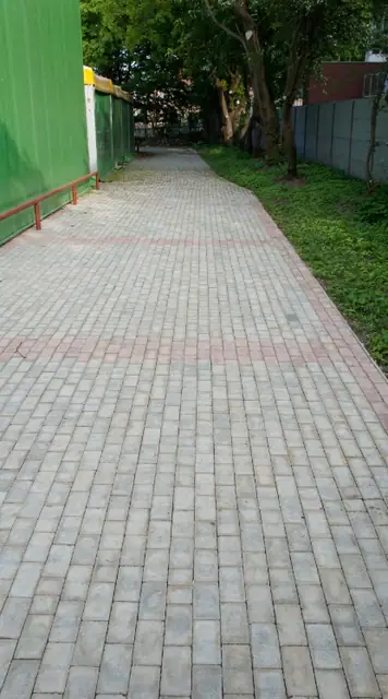 Chodnik z szarej kostki brukowej prowadzący wzdłuż zielonego ogrodzenia i trawnika